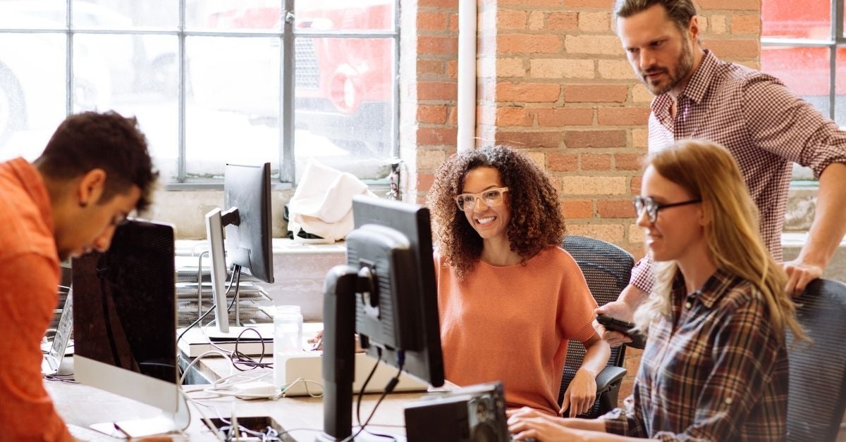 Four colleagues collaborating around a shared desk with computers in a bright, modern office with an exposed brick wall.