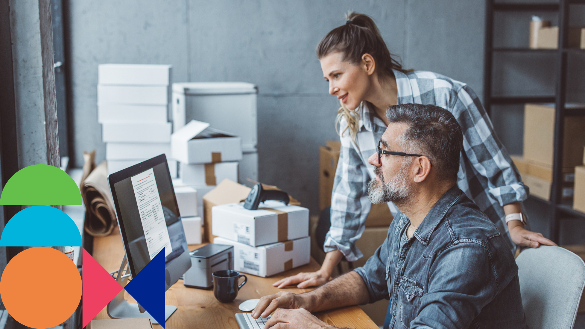 A man and a woman in a small business office looking at a desktop computer monitor next to stacked shipping boxes.